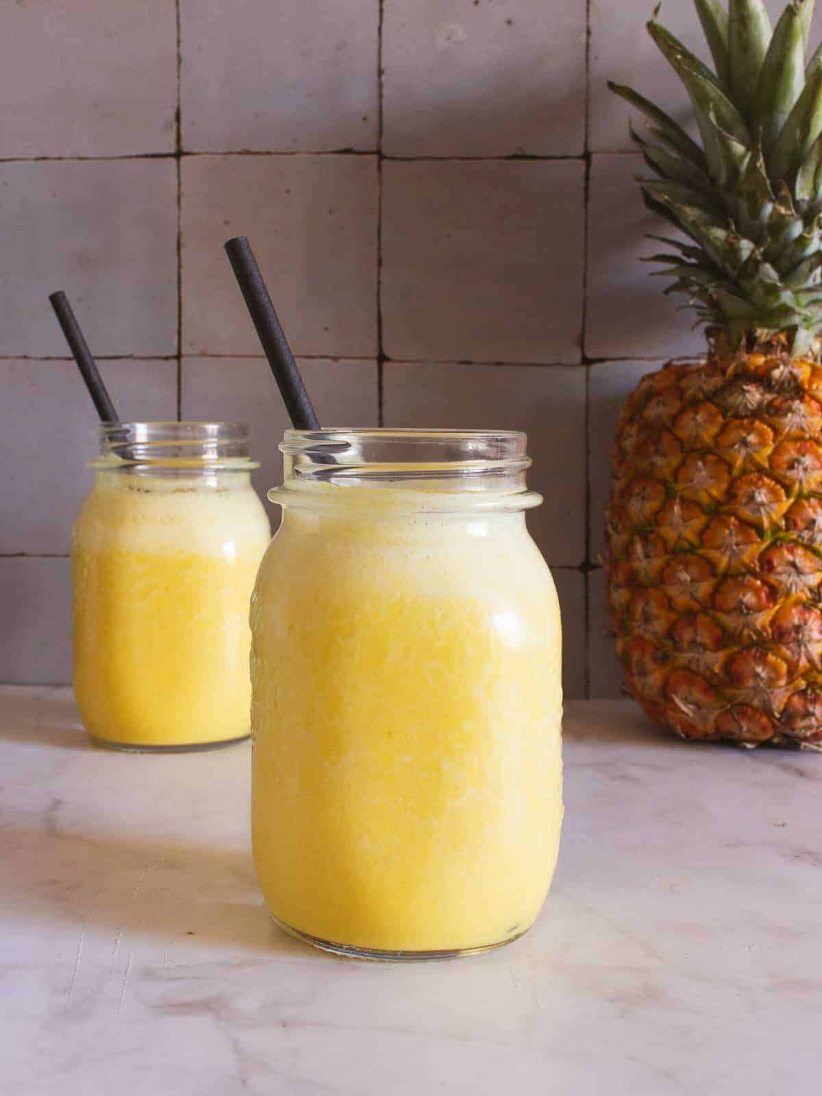 two Pineapple Coconut Smoothie glasses in a marble table with a whole pineapple behind