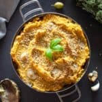 Cazuela con polenta al horno crocante y dorada, decorada con hojas de albahaca fresca, sobre una mesa oscura con ajo, orégano y una cuchara con restos de polenta a su alrededor.