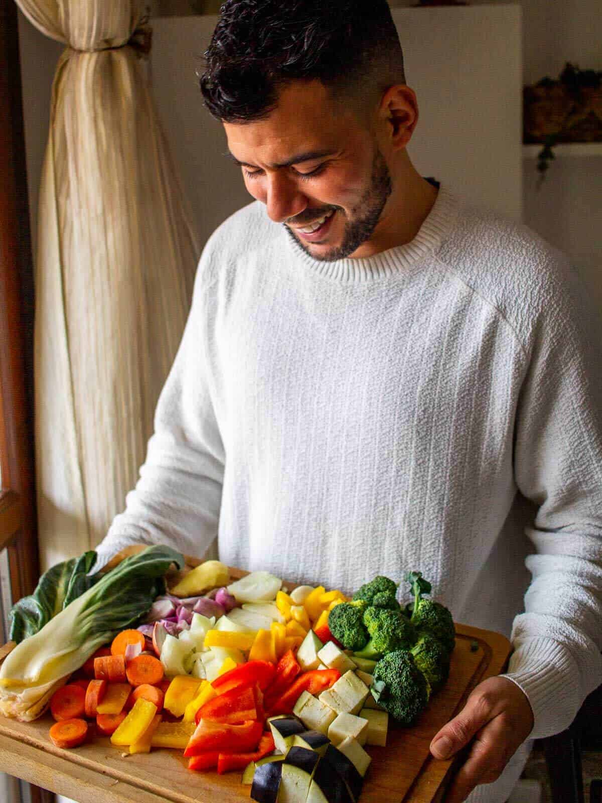 Hombre sosteniendo una tabla de madera llena de verduras frescas y coloridas como brócoli, zanahoria, pimiento, berenjena junto a una ventana con luz natural.