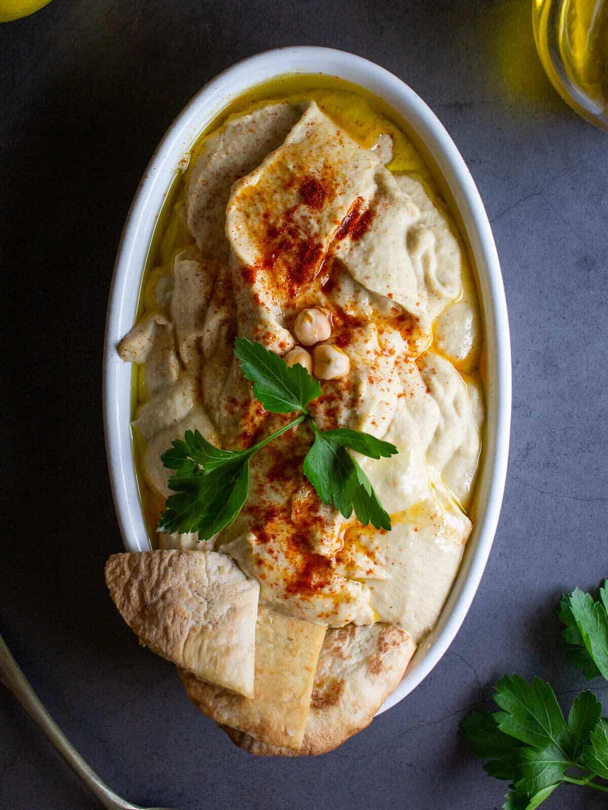 hummus clásico servido en un plato ovalado adornado con pimentón, chips de pita y perejil plano.