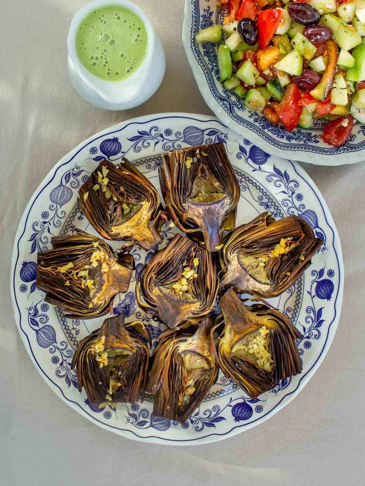 plato con alcachofas al horno junto a una jarrita con salsa verde y un plato con ensalada griega.