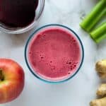 vaso servido con jugo de betabel junto a una manzana, un trocito de jengibre, apio y betabel.