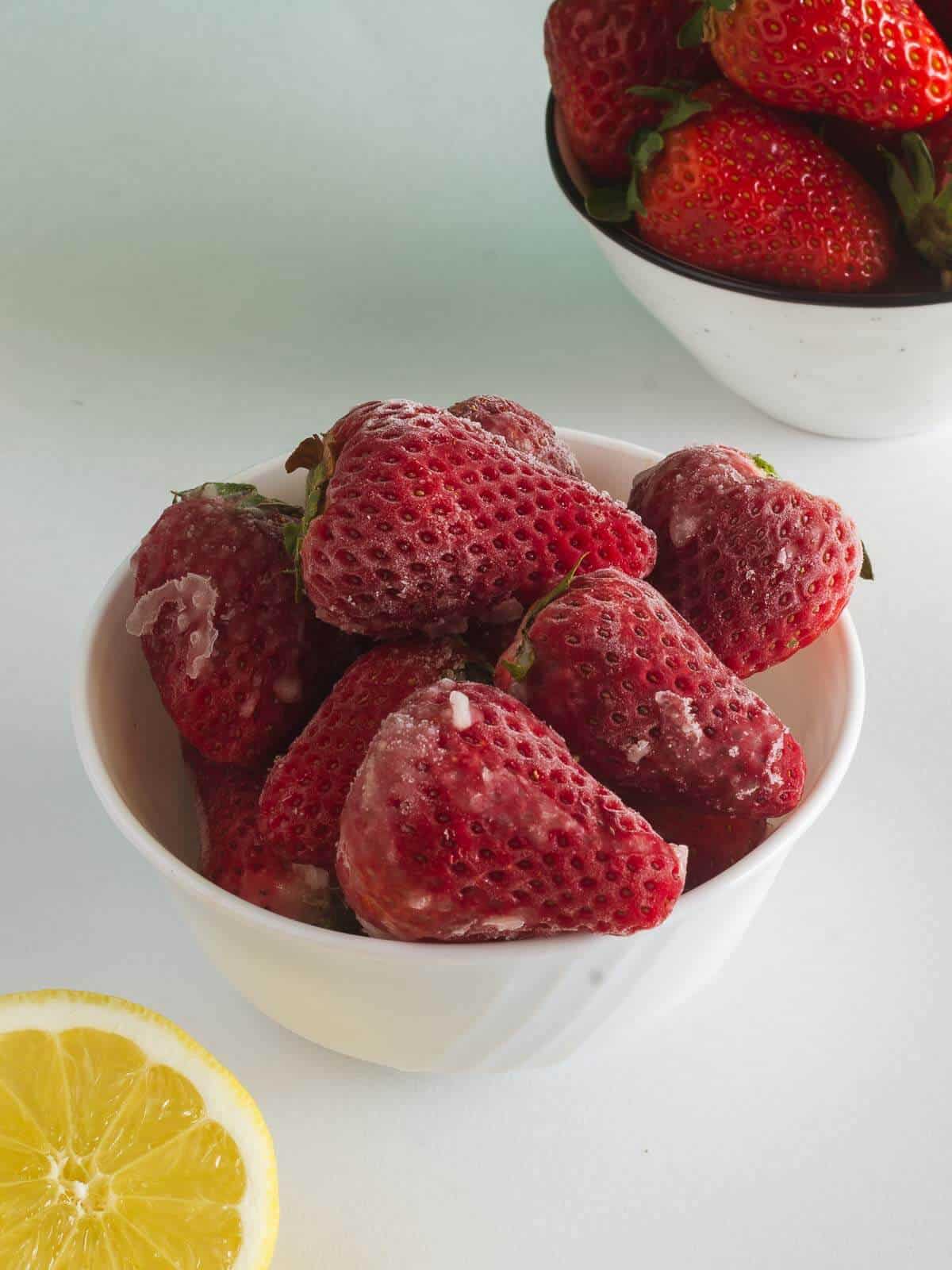 un bowl con fresas congeladas junto a otro con fresas frescas y medio limón.