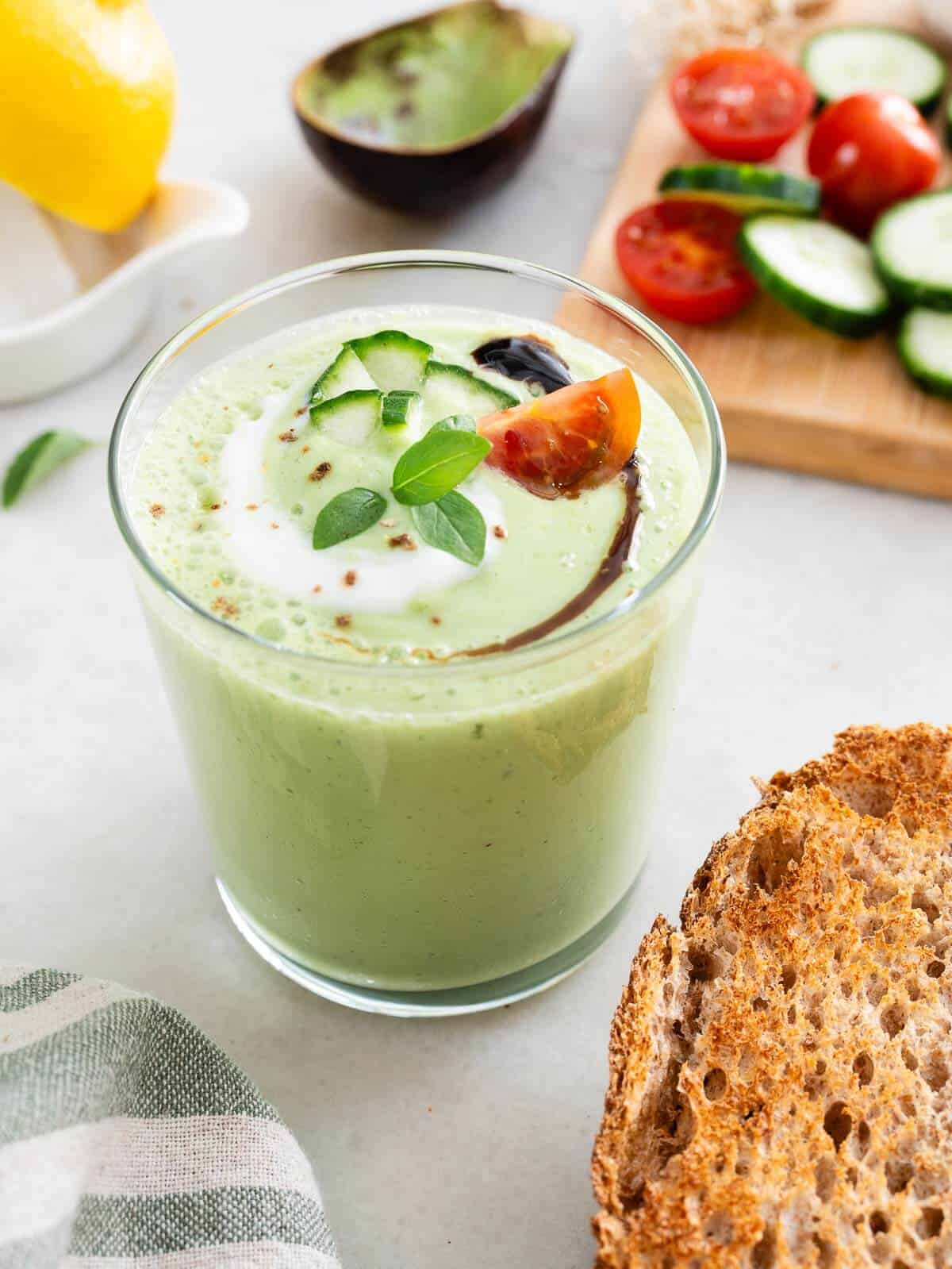 crema de pepino y aguacate servida en un vaso, junto a una rodaja de pan tostado y una tabla con mitades de tomates y rodajas de pepino.