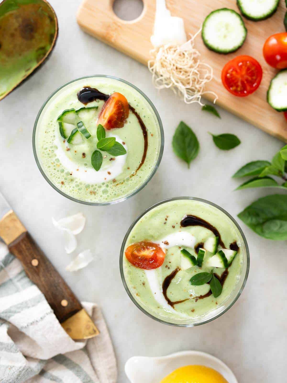 dos vasos de crema de pepino y aguacate decorados con un remolino de glaseado balsámico, rodajas de tomate, pepino y yogur.