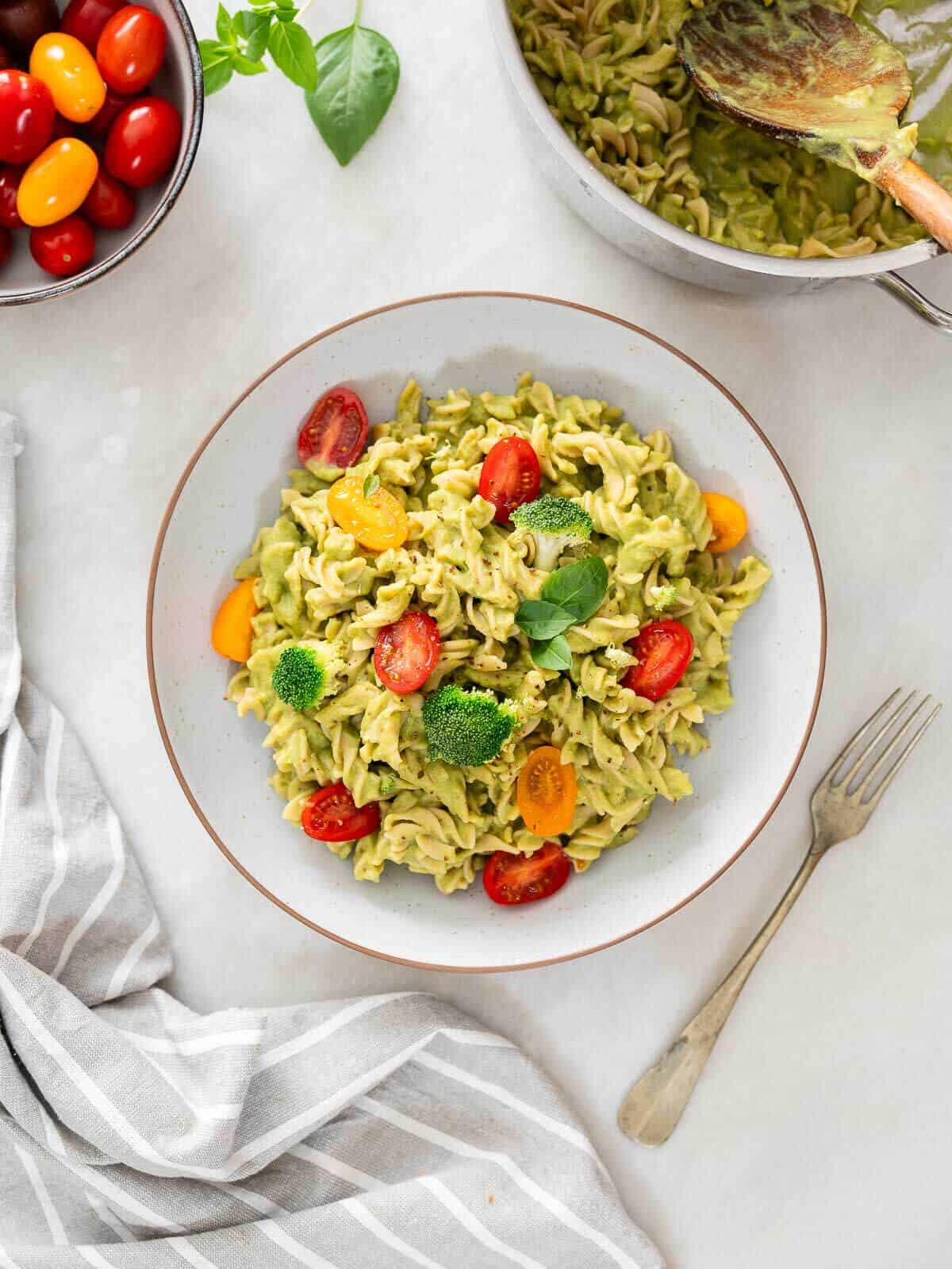 bol de ensalada de pasta verde saludable rica en proteínas, adornada con tomates cherry cortados por la mitad, brócoli y hojas de albahaca fresca, acompañada de un bol de tomates cherry y un paño de cocina