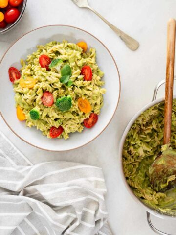 bol de ensalada de pasta con brócoli cremosa con una cuchara de madera, mostrando la textura cremosa y la mezcla de verduras, con una sartén con más ensalada de pasta visible en el fondo.