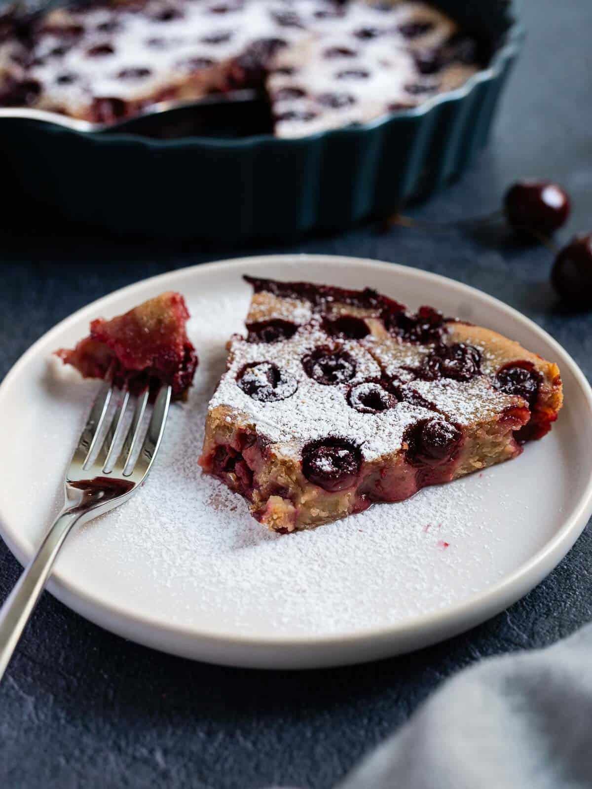 Tarta de cerezas estilo clafoutis en un plato, servida con un tenedor, espolvoreada con azúcar glas.