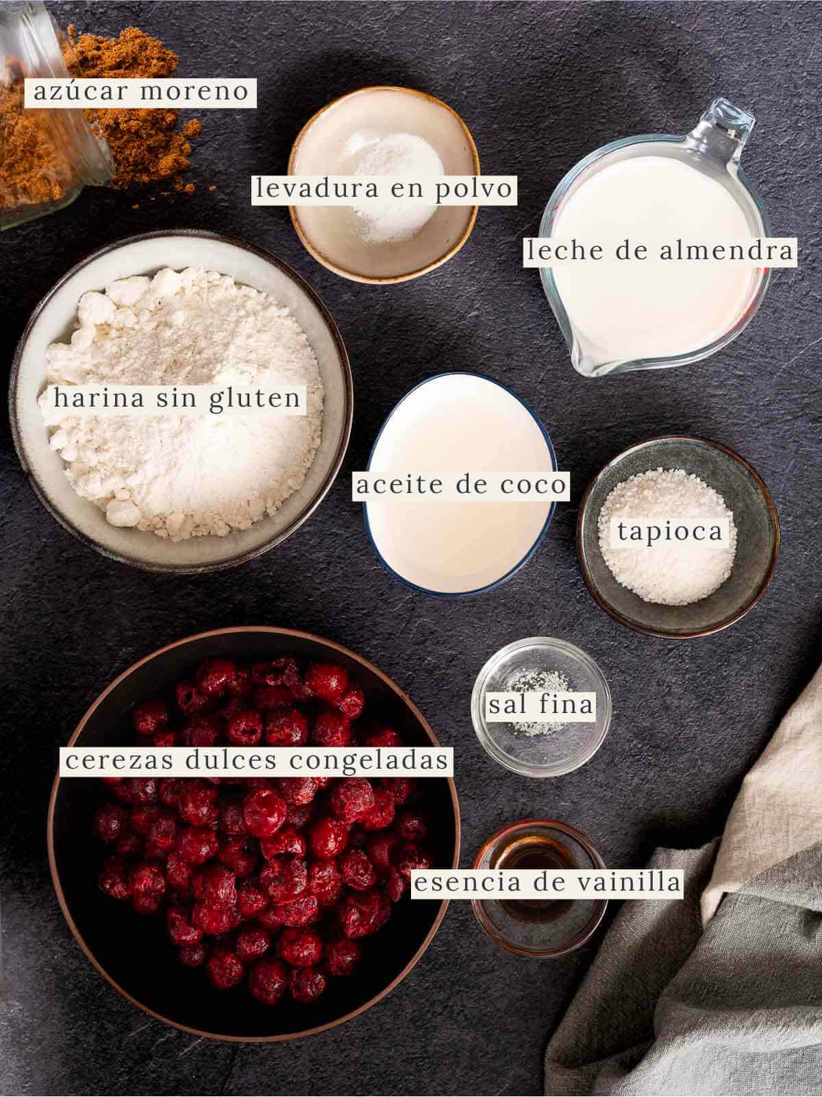 Ingredientes de la tarta de cerezas organizados: harina sin gluten, leche de almendras, aceite de coco, azúcar moreno, tapioca, cerezas congeladas, esencia de vainilla, levadura en polvo y sal fina.