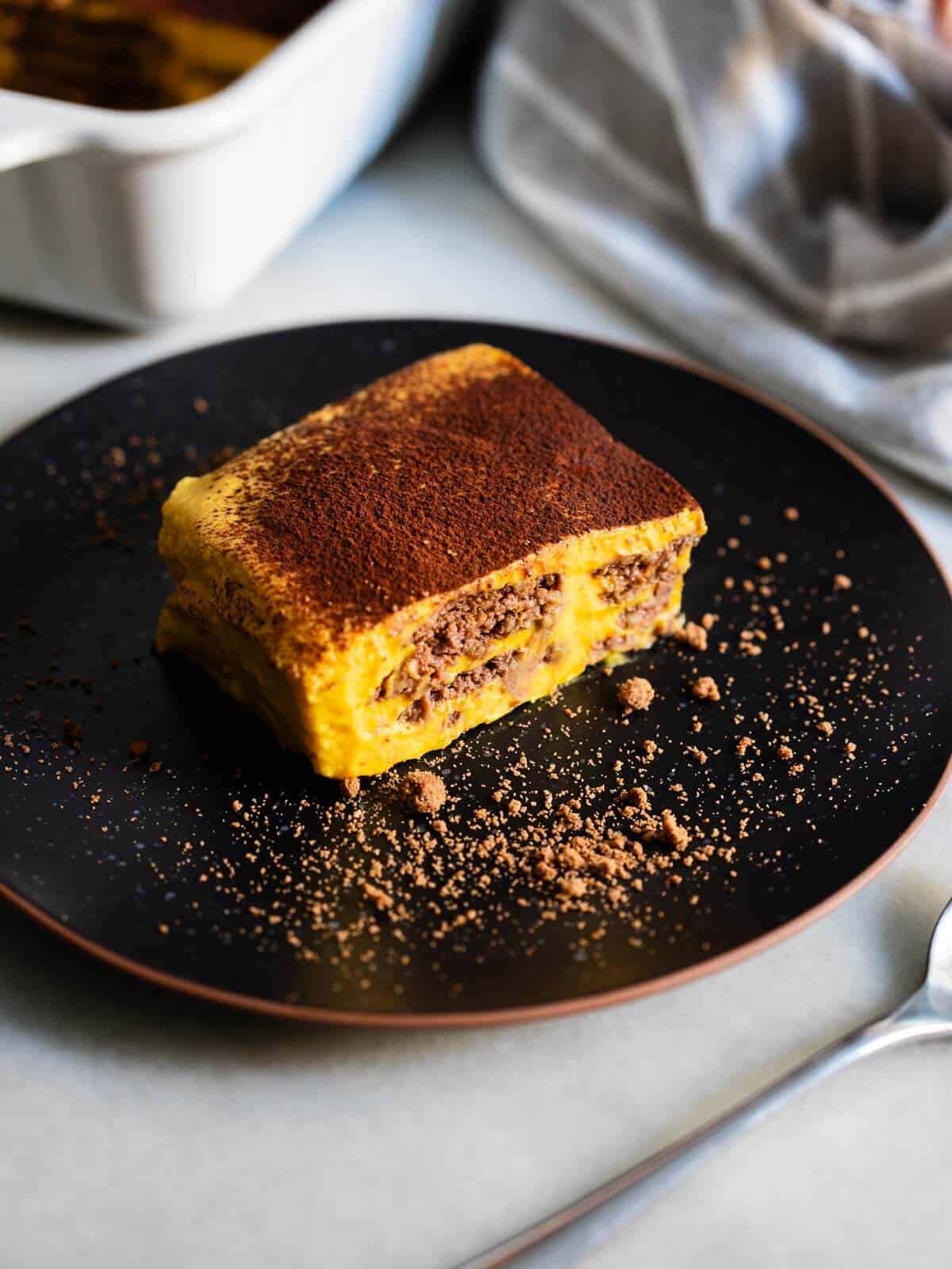 Porción de tarta de galletas de chocolate con calabaza en un plato negro, con cacao en polvo espolvoreado alrededor.