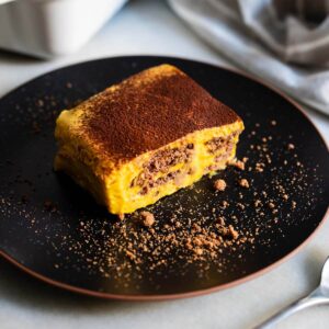 Porción de tarta de galletas de chocolate con calabaza servida en un plato negro, espolvoreada con cacao en polvo.