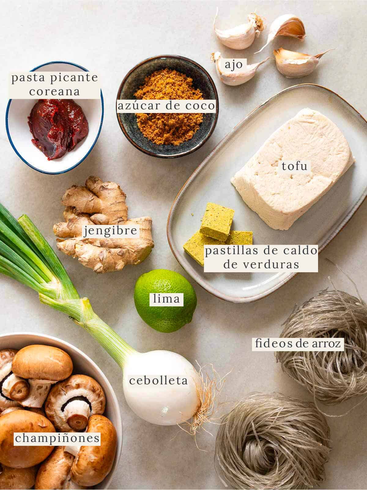 Vista cenital de los ingredientes de la sopa picante de tofu coreano, que incluye tofu, ajo, lima, champiñones cremini y fideos de camote, dispuestos de manera ordenada.