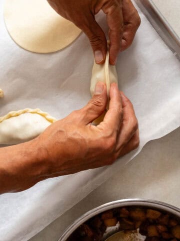 Primer plano de dos manos doblando un disco de empanada alrededor del relleno de manzana, con otras empanadas alineadas en una bandeja, listas para ser selladas.