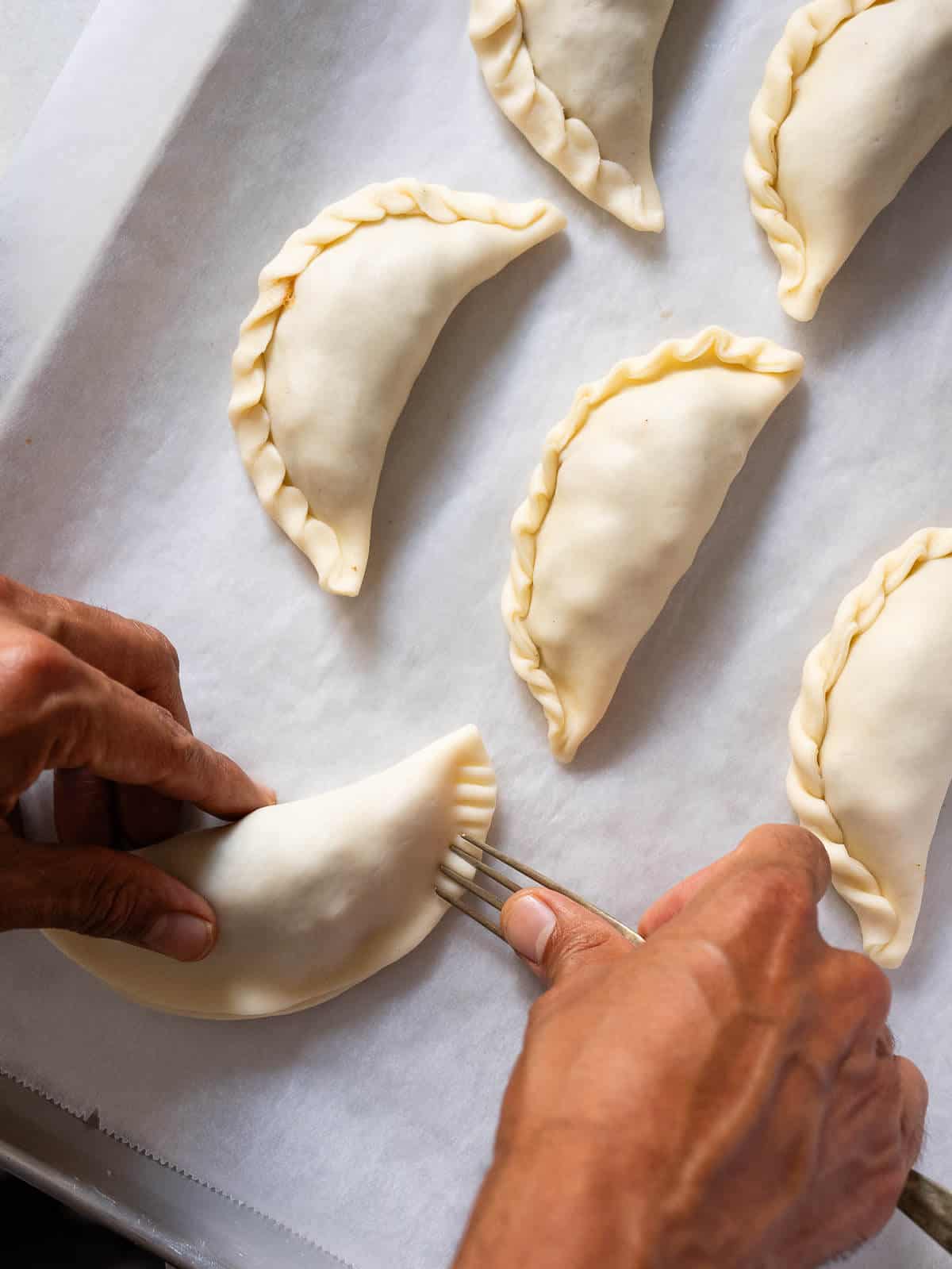 Bandeja de horno con papel encerado con empanadas de manzana colocadas en filas, cerrando una de ellas con la punta de un tenerdor.