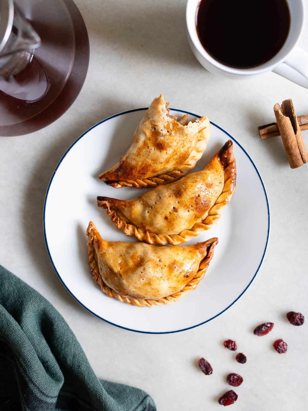 Un plato con dos empanadas de manzana recién horneadas, doradas y ligeramente crujientes, con un toque de arándanos secos al fondo.