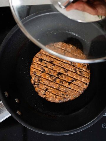 Hamburguesas de frijoles negros cocinándose en una sartén bajo una tapa transparente.