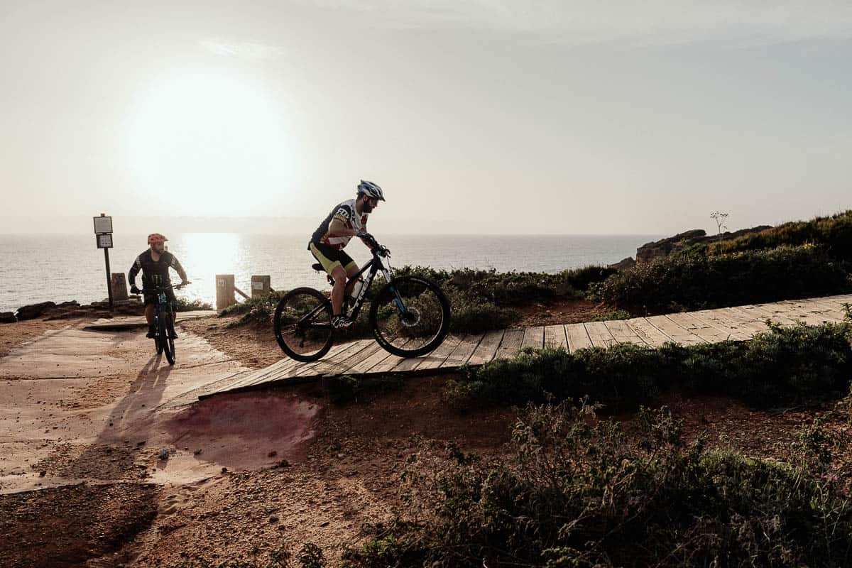 Dos ciclistas recorren un sendero pintoresco cerca del borde de un acantilado, con el sol poniéndose en el fondo. La luz dorada del atardecer arroja un brillo cálido sobre el paisaje, destacando el terreno accidentado y el vasto océano abajo.