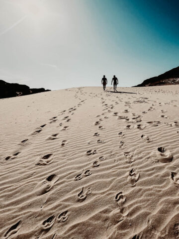 Dos personas caminan a través de vastas dunas de arena bajo un cielo brillante, sus huellas creando un rastro detrás de ellas. El sol proyecta un efecto dramático de luz y sombra sobre las dunas, destacando la textura de la arena y el sentido de aventura en este entorno natural.