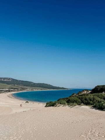 Vista impresionante de la Playa de Bolonia con su extensa costa de arena que se encuentra con el mar azul claro. La playa está rodeada de vegetación verde y colinas distantes, ofreciendo un paisaje costero pintoresco y tranquilo.