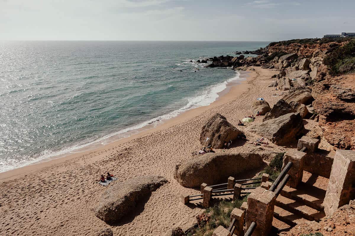 Vista aérea de las Calas de Roche, una playa aislada rodeada de dramáticos acantilados rocosos y aguas cristalinas de color turquesa. La orilla arenosa está salpicada de grandes rocas y unos pocos bañistas, disfrutando del entorno tranquilo y pacífico. Las escaleras que descienden a la playa añaden un toque de encanto rústico a este pintoresco lugar costero.