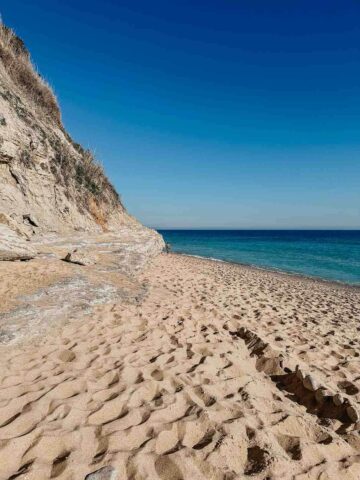 Vista de una playa de arena virgen en Caños de Meca con acantilados dramáticos a la izquierda y aguas claras y azules que se extienden hacia el horizonte bajo un cielo azul brillante.