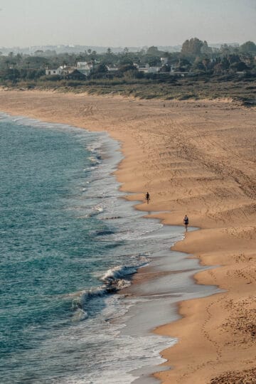 Un largo tramo de playa en la Playa Faro de Trafalgar, con suaves olas que rompen en la orilla y algunas personas caminando por la orilla.