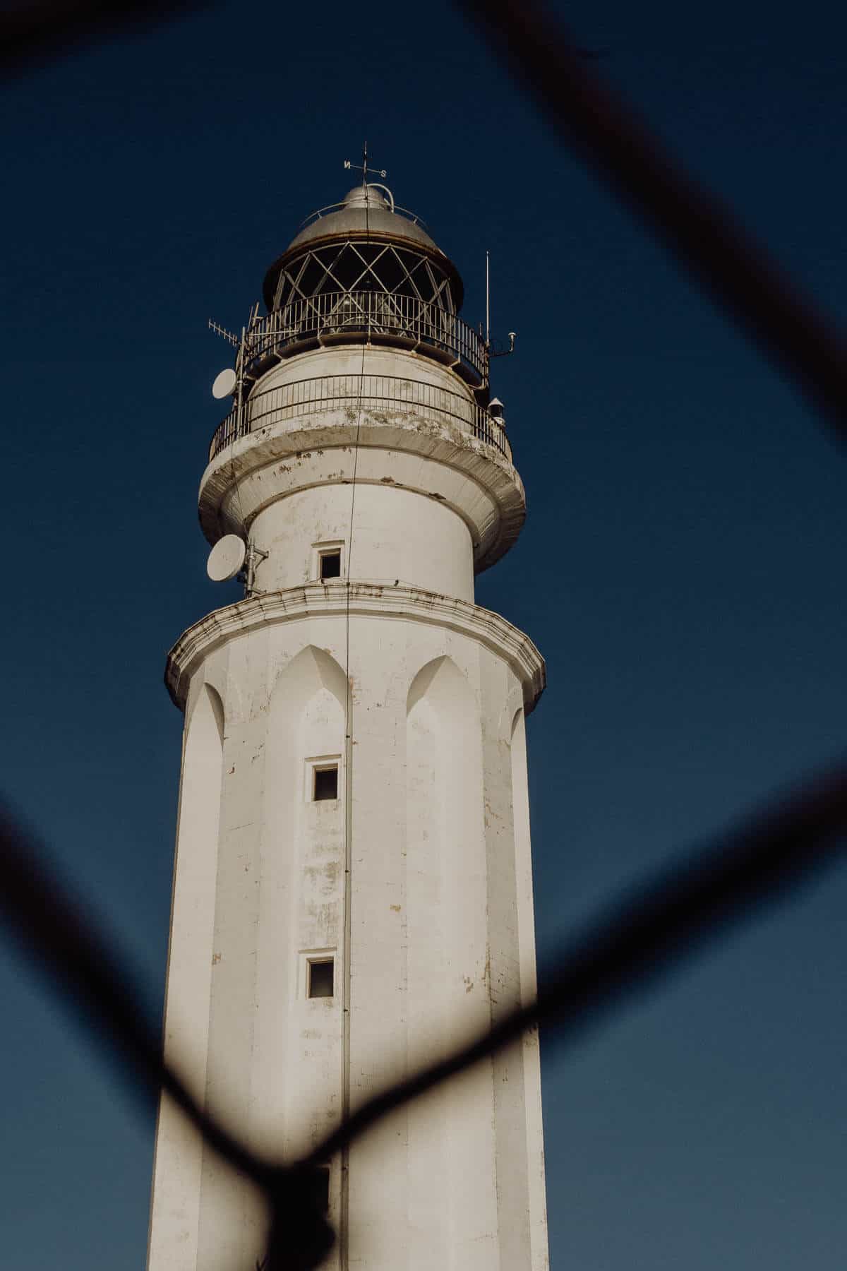 Una vista de cerca de un histórico faro blanco contra un cielo azul profundo, enmarcado por un elemento oscuro en primer plano desenfocado.