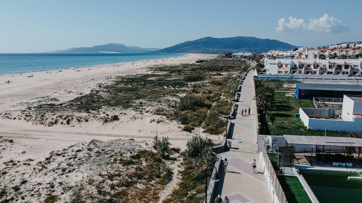 Vista aérea de El Palmar de Vejer, mostrando una larga playa de arena que se extiende a lo largo de la costa, con una mezcla de vegetación natural y espacios abiertos. A la derecha, un paseo pavimentado corre paralelo a la playa, bordeado de edificios modernos y áreas residenciales. En la distancia, montañas y un cielo azul claro proporcionan un telón de fondo pintoresco.