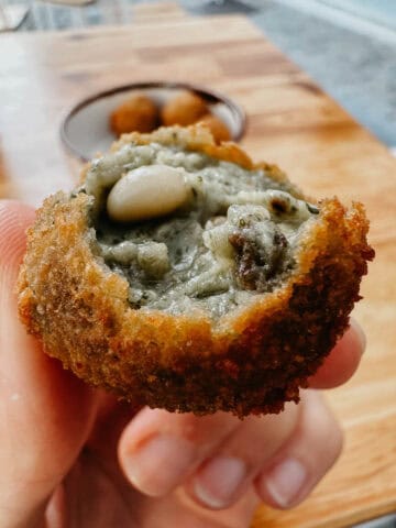 Primer plano de una mano sosteniendo una croqueta rellena de setas y piñones, mostrando su crujiente exterior y su sabroso relleno.