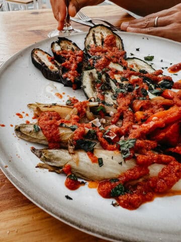 Un plato de verduras a la parrilla cubiertas con una salsa de tomate espesa en El Lola. El plato incluye berenjena, calabacín y otras verduras, adornadas con hierbas, mostrando una opción vegana vibrante y apetecible.