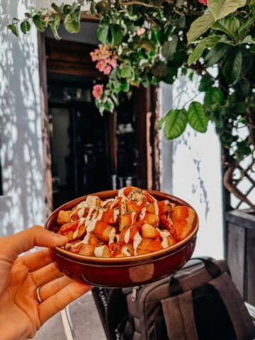 Una mano sosteniendo un cuenco de patatas bravas cargadas, cubiertas con salsas y hierbas. El fondo muestra la entrada de Bar el Francés, adornada con plantas y flores verdes, proporcionando un ambiente acogedor e invitador.