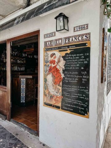 La entrada de Bar el Francés, con un encantador cartel vintage y un menú en pizarra que muestra varios platos. El exterior está decorado con plantas y flores, lo que añade al ambiente pintoresco y acogedor del restaurante.