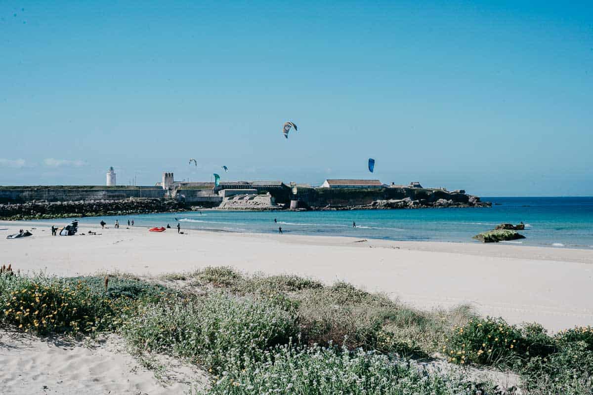 Una vista panorámica de una playa en Cádiz con arena dorada, aguas cristalinas y kitesurfistas en la distancia. La costa está enmarcada por vegetación en primer plano y una fortaleza histórica y un faro al otro lado de la bahía.