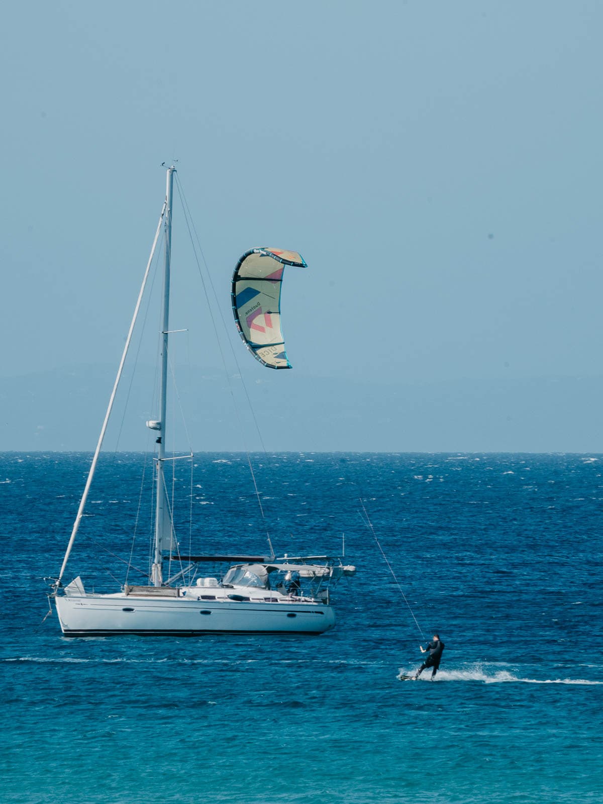 Un velero está anclado en el mar tranquilo con un kitesurfista deslizándose cerca, creando una escena serena y aventurera contra las claras aguas azules.