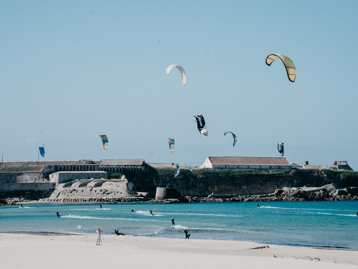 Una escena costera muestra varios kitesurfistas deslizándose sobre el agua, sus coloridas cometas surcando el cielo. Al fondo, una fortificación histórica se encuentra en la costa rocosa, contrastando con el mar azul brillante y el cielo despejado.