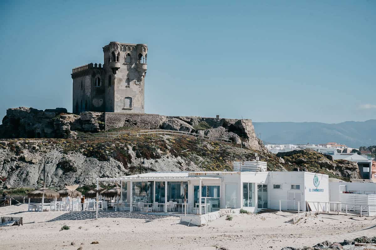 Una escena de playa con una torre de piedra histórica en lo alto de una colina rocosa, con vista a un moderno restaurante de playa llamado "El Chiringuito". El restaurante tiene un diseño blanco y minimalista con áreas de comedor al aire libre, contrastando con la antigua arquitectura de la torre.