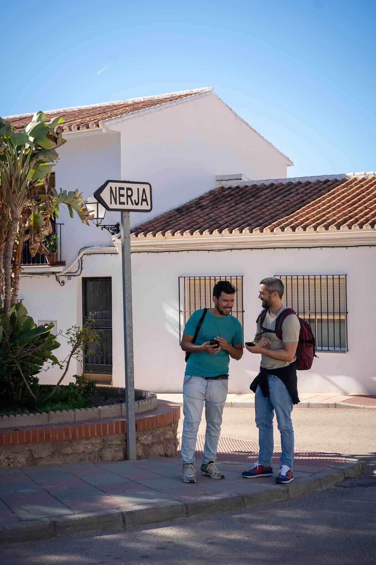 Dos chicos hablando en la calle junto a un cartel con "Nerja" escrito.