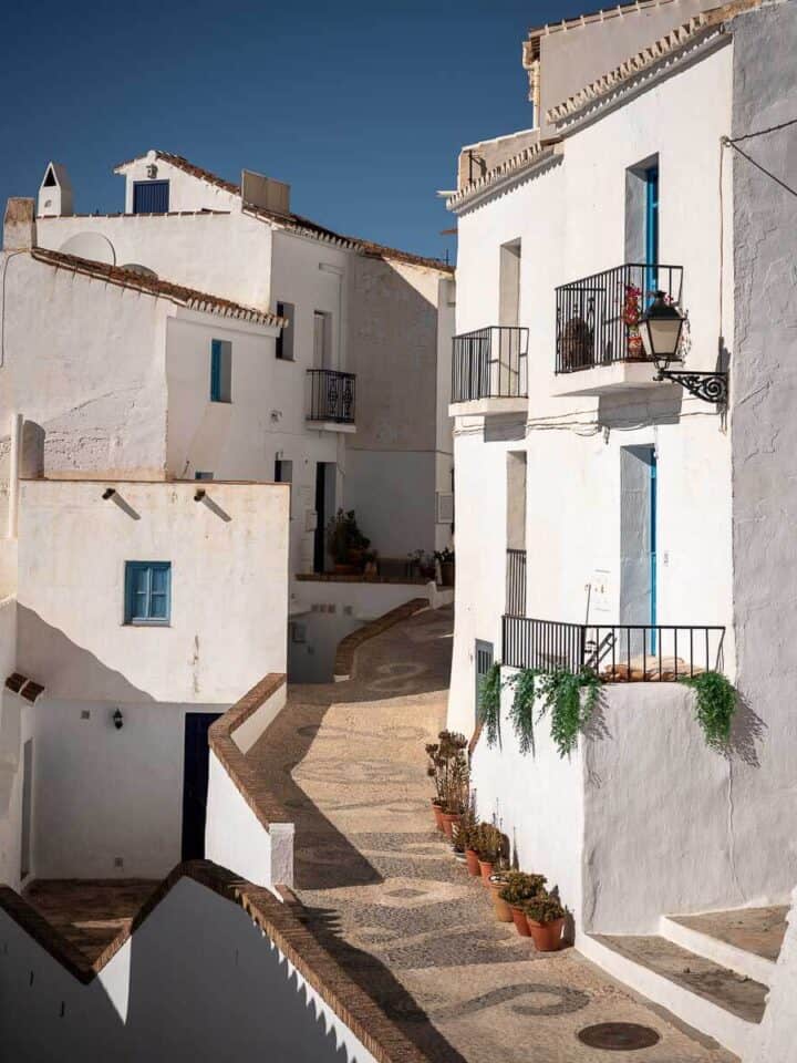 Calle empedrada con casas encaladas de estilo andaluz en Frigiliana, con detalles en azul en ventanas y balcones.