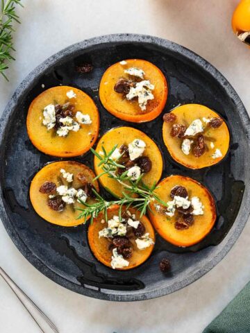 Rodajas de caqui persimón servidas con queso azul, pasas hidratadas y decoradas con hojas frescas de romero en un plato oscuro.