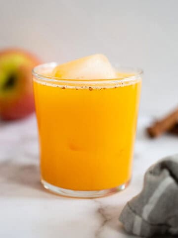 Vaso de jugo de calabaza y manzana servido con un cubo de hielo, con manzanas frescas y canela en el fondo. Bebida ideal para los días de otoño en busca de sabores auténticos y saludables.