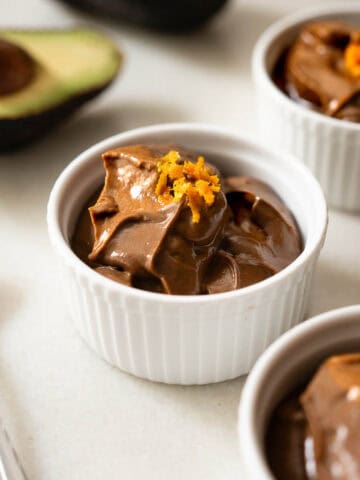 Mousse de chocolate con aguacate servido en ramequines blancos, decorado con ralladura de naranja, postre vegano cremoso y saludable.