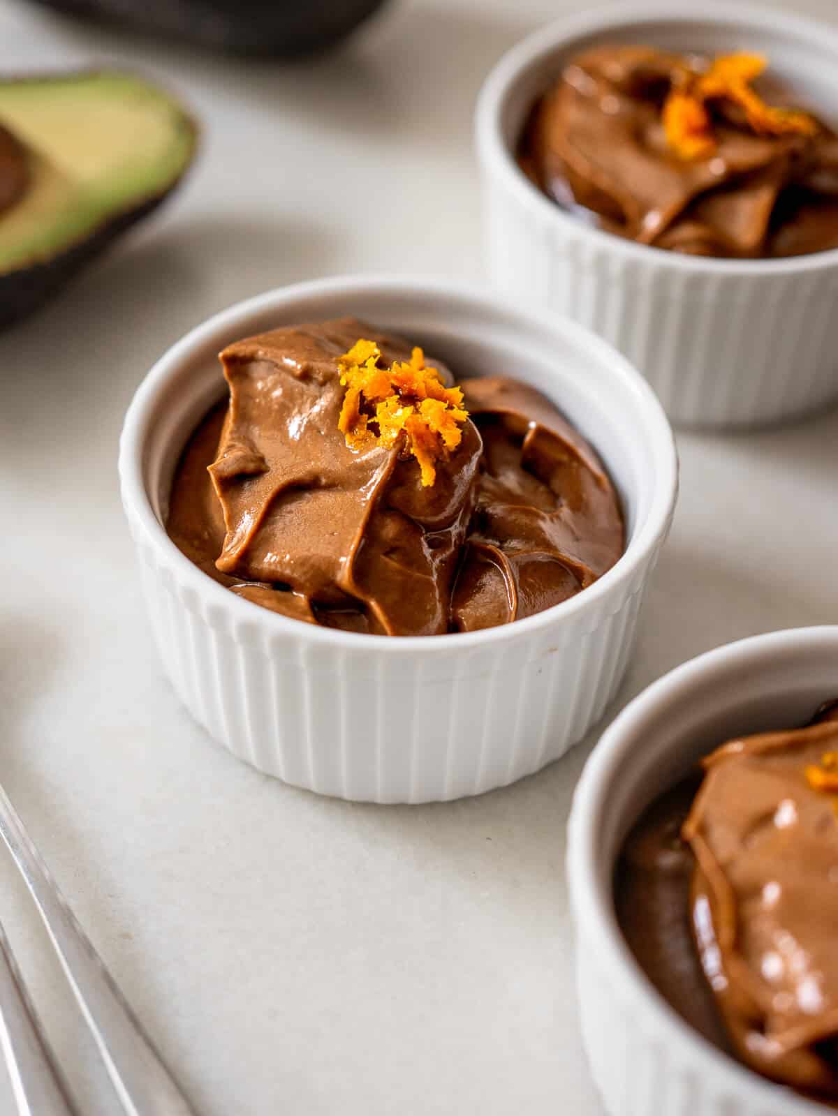 Mousse de aguacate y cacao servido en ramequines blancos con ralladura de naranja, opción de postre saludable.