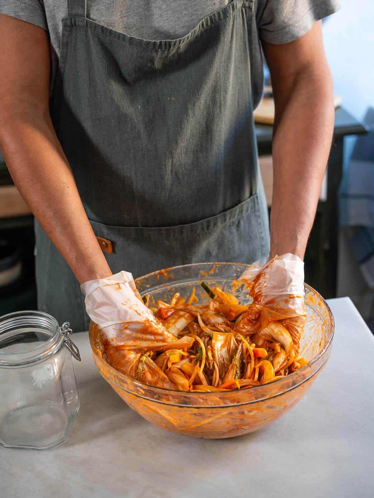 Manos mezclando col china y vegetales con pasta de gochugaru en un bol grande. Paso clave en la preparación del kimchi vegano casero fermentado al estilo coreano.