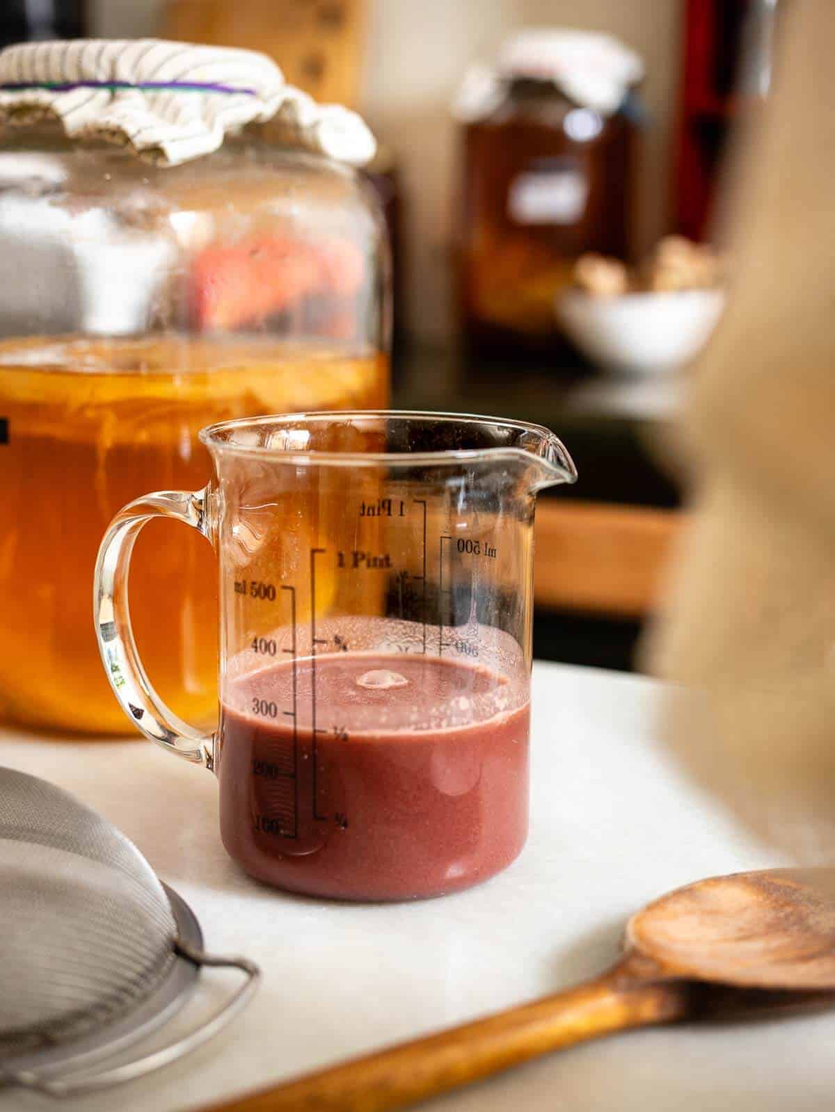 Vaso medidor con pulpa de frutas listo para la segunda fermentación de kombucha, con el frasco de fermentación en el fondo.