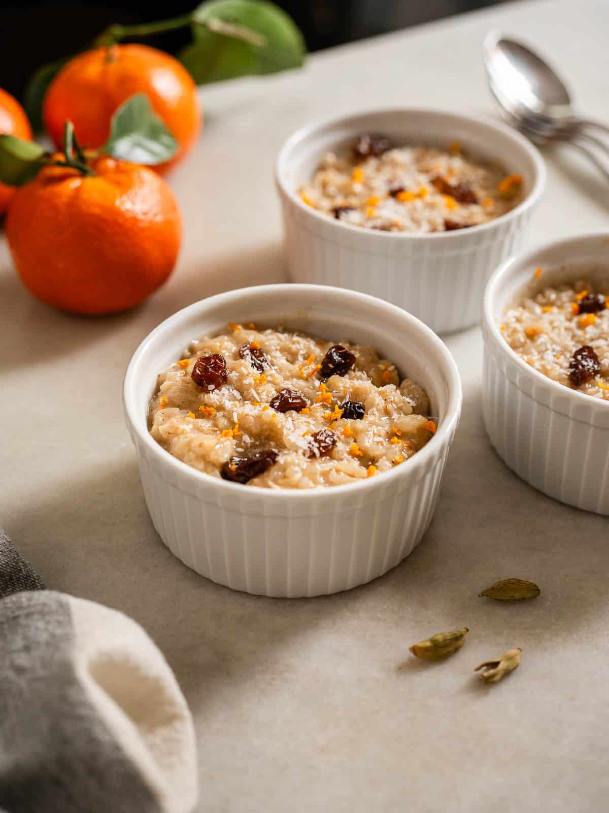 Tres porciones de postre de arroz cremoso en ramequines blancos con pasas y especias, con mandarinas de fondo.