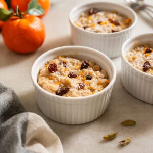 Dos ramequines con postre de arroz cremoso al cardamomo, acompañados de mandarinas frescas y semillas de cardamomo sobre una mesa.