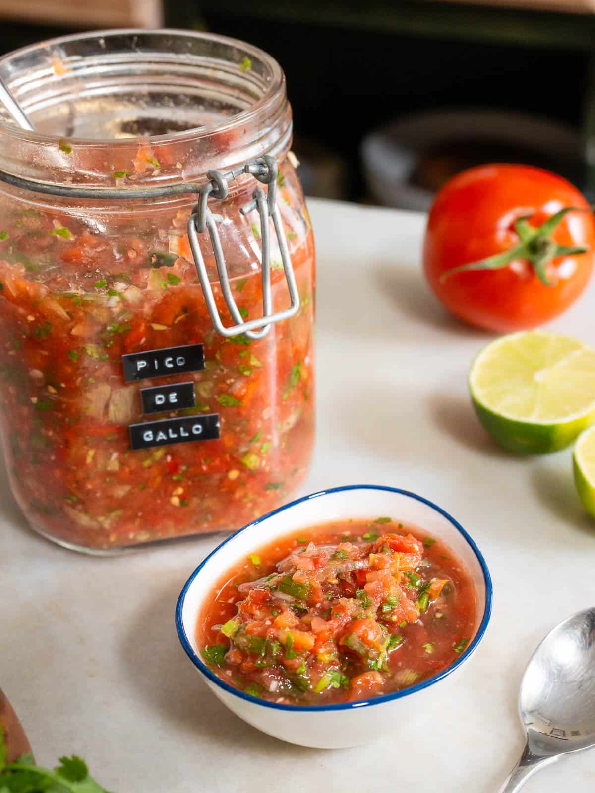 Tazón pequeño con pico de gallo casero junto al frasco lleno, rodeado de limas y tomates frescos.