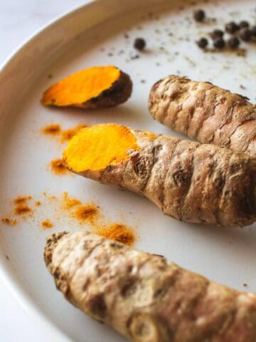 Raíz de cúrcuma fresca cortada mostrando su interior anaranjado, acompañada de pimienta negra en un plato blanco.