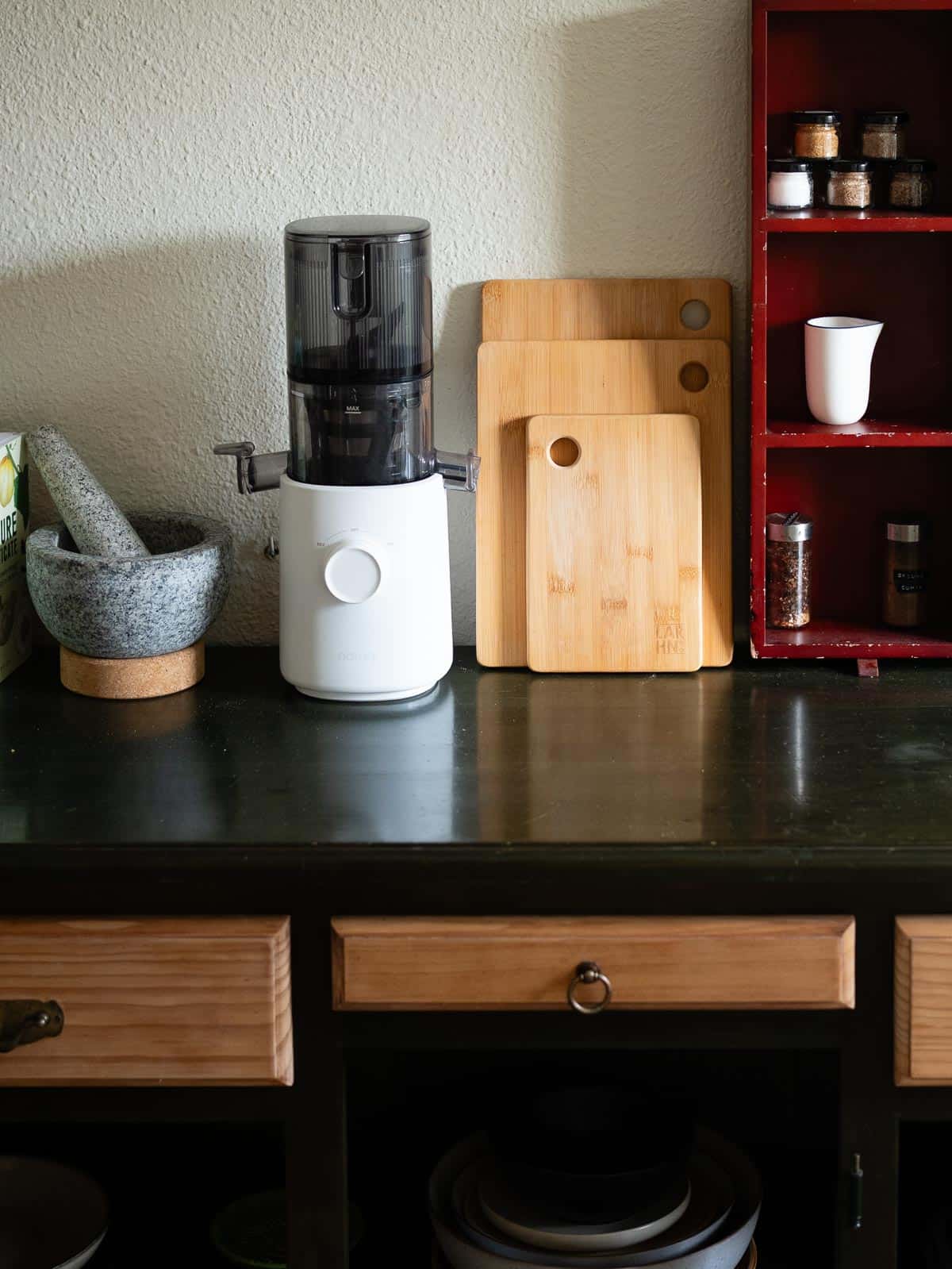 Superficie de cocina con tablas de cortar de bambú apoyadas contra la pared, junto a un extractor de jugos blanco, un mortero de granito y un estante rojo con frascos de especias.