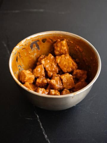 Tempeh en cubos cubierto con salsa de cacahuete y sriracha en un bol metálico.